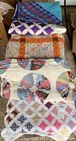 Five handmade vintage quilts and quilt tops stacked showing various patchwork patterns and colors including purple, white, orange, multicolor, blue, and teal.