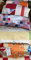 Overview of five stacked vintage handmade quilts and quilt tops showing various patterns and colors including star and patchwork designs.