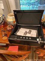 Open black Sentry Safe showing key lock, fingerprint scanner, keypad buttons, two keys on a ring, and owner's manual inside the compartment.