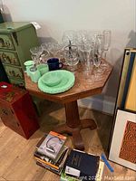 Hexagon shaped side table with wooden pedestal base, displaying multiple glassware items including wine glasses, champagne flutes, goblets, and colored dishware on top.