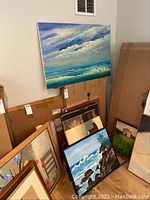 Group shot of nine artworks stacked and leaning against a wall, showing variety of sizes, frames, and subjects including ocean scene with sailboat and waves.