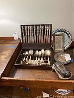 Wide photo showing entire flatware set in box with knives on lid, forks and spoons in bottom, alongside silver-toned serving dishes including butter dish and plates.