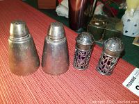 Two pairs of salt and pepper shakers on a red surface: one plain tapered pair, one pair with filigree outer shells and glass inner containers