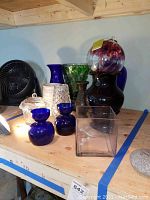 Wide shot showing all vases and decorative glass items arranged on wooden table: includes cobalt blue vases, clear glass vase, green floral vase, dark purple vase, glass ball, crystal cut vase, and glass cupcake container.