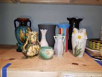 View of all nine vases arranged on a shelf showcasing variety in color, style, and size including majolica and floral designs.