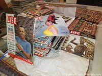 Photo showing stacks of LIFE magazines with some visible covers including a man in blue on May 19, 1961 cover and a woman in yellow with a red hat.