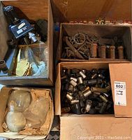 Top view of a box containing a vintage microscope head, multiple boxes with metal parts, hardware, and packaged glass lenses.