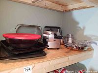 Shelf with Bialetti waffle maker, glass mixing bowl with red lid, metal cake pan, metal strainer, and other bakeware items.