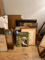 Photo showing a group of wall art pieces leaning against a wall, including framed portraits and landscape paintings, some unframed and a large double-sided piece.