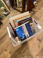 Photo of a translucent plastic tote full of various small sized artworks on boards or canvas, some framed and some unframed, standing upright.