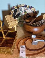 Photo showing wooden cutlery block with knives, two wooden bowls (one striped, one plain), woven basket, decorative tray with floral and geometric patterns, and a small grater with orange base.