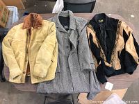 Three vintage jackets on table including tan suede McCloud style jacket, black and beige suede bomber, and herringbone tweed overcoat.