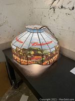 Image of the Budweiser Tiffany hanging lamp set on a black table, displaying the stained glass design featuring Budweiser branding and Clydesdale horses.