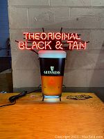 Lit neon sign displaying 'THE ORIGINAL BLACK & TAN' text and pint glass image with Guinness and Bass logos, mounted on a wooden surface against a cinder block wall.