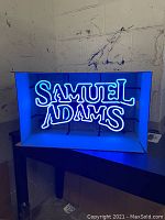 Lit neon sign showing 'SAMUEL ADAMS' in bright blue light on a black backing.