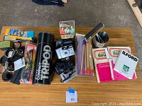 Full lot overview with cameras, tripod container, flash cubes, film rolls, and darkroom supplies spread on a table.