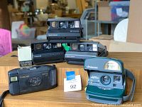 Front view of Polaroid Spectra System, One Step, Captiva SLR and Self Timer cameras, arranged on table with some accessories box.