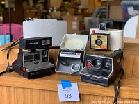 Overview photo showing all five Polaroid cameras and accessories arranged on a wooden surface including the Polaroid Sun 600, Land Camera 103, Close-Up Attachment, Land Camera Swinger, and Polaroid Pronto.