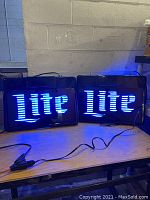 Two Miller Lite neon signs illuminated, showing blue neon "Lite" text. Power cords visible on table.