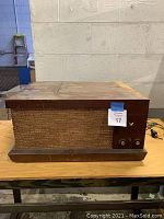 Front of vintage phonograph showing wooden cabinet with woven fabric speaker grille, two control knobs and toggle switch, and V-M logo.