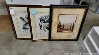 Three framed artworks leaning on the floor: two botanical prints with white flowers and one landscape painting with decorative mat