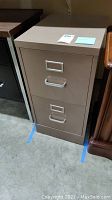 Side front view of the closed two drawer metal file cabinet in brown color with silver handles and label holders.