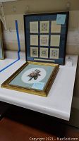 Overall view of two framed pieces displayed side by side: one blue mat frame with nine art prints, one gold frame with floral petit point needlework.