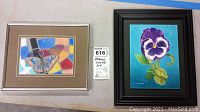Two framed colored pencil art pieces on a grey surface. Left artwork shows butterfly with colorful geometric background, right artwork shows purple pansy flower. Both signed M. Kaestner.