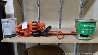 Black & Decker hedge trimmer model BEHT150 with instruction manual next to Scotts Whirl hand-powered spreader on shelf.