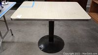 Full view of rectangular kitchen table with laminated wood top and iron pedestal leg standing on concrete floor