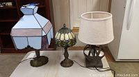 Three table lamps on floor: two with stained glass shades and brass-like bases; third with modern metal base and fabric shade.