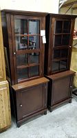 Two matching dark wood display cabinets with glass-paneled upper doors and lighted adjustable shelves, lower wooden doors for storage, standing side by side.