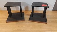 Two black vintage Plateau speaker stands side by side showing top platform and walnut accented base on wood floor.