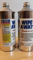 Photo showing two metal containers of Wipe Away remover with health and safety hazard labels on side and product information.