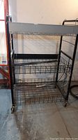 Front view of black metal rolling cart with three wire basket shelves and caster wheels on concrete floor.