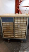 Front view showing two columns of small drawers, most with paper labels, one larger blue drawer, four casters at bottoms for mobility.