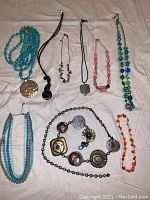 Eight necklaces displayed flat on fabric surface alongside seven disk metal belt