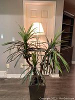 Faux plant with multiple long, narrow-leaved stems packed densely, placed in a tall black square resin planter with decorative stones at the base. Plant approximately 40x60 inches.