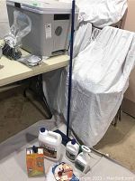 Photo showing Bona hardwood floor cleaner bottles, mop, and Swiffer duster arranged on floor and against covered chair.