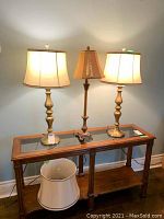 Two bronze gold metal tri-light lamps with creamy sateen fabric lampshades on a wooden table with glass top; one ornate brown gold lamp with tasselled shade between them.