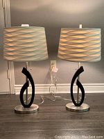 Two identical modern table lamps shown side by side on dark wooden floor. Each lamp has a white textured cotton drum shade and black curved midsection on brushed nickel base. Silver translucent cords visible.