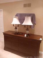 Pair of matching table lamps on a wooden dresser against a wall with fabric covering behind.