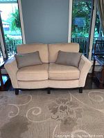 Front view of the light creamy brown love seat with two rectangular pillows on a beige patterned rug.
