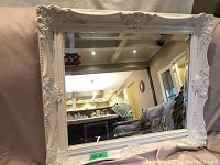 Front view of rectangular white wood frame mirror with ornate corner carvings and beveled glass, reflecting interior ceiling and walls.