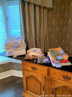 Linen and napkins arranged on wood cabinet near window with decorative drapes, showing various folded white linens and a small bag of napkin rings