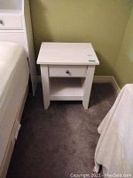 Side angled view of one white melamine nightstand showing drawer, open shelf, and part of the surrounding area