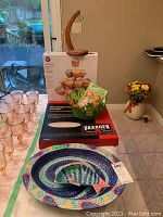 Overview of the lot items including new boxed Wilton cupcake stand, wooden banana tree stand, ceramic pizza stone and rack box, felt butterfly flower clip package, pink glass dessert cups, mosaic fish platter and dip dish.