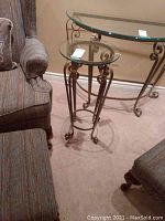 Full view of the wrought iron and glass plant stand next to upholstered chairs, showing round glass top and iron scroll detail.