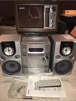 Sony Micro Hi-Fi stereo main unit, two speakers, remote control, and owner's manual laid out on table with vintage Sony radio on top