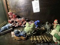 First shelf view showing red carved stone sculpture, green jade-like chicken and chick, blue speckled cat on wooden base, pink porcelain cat, set of brass cats, and deep blue stone figurine